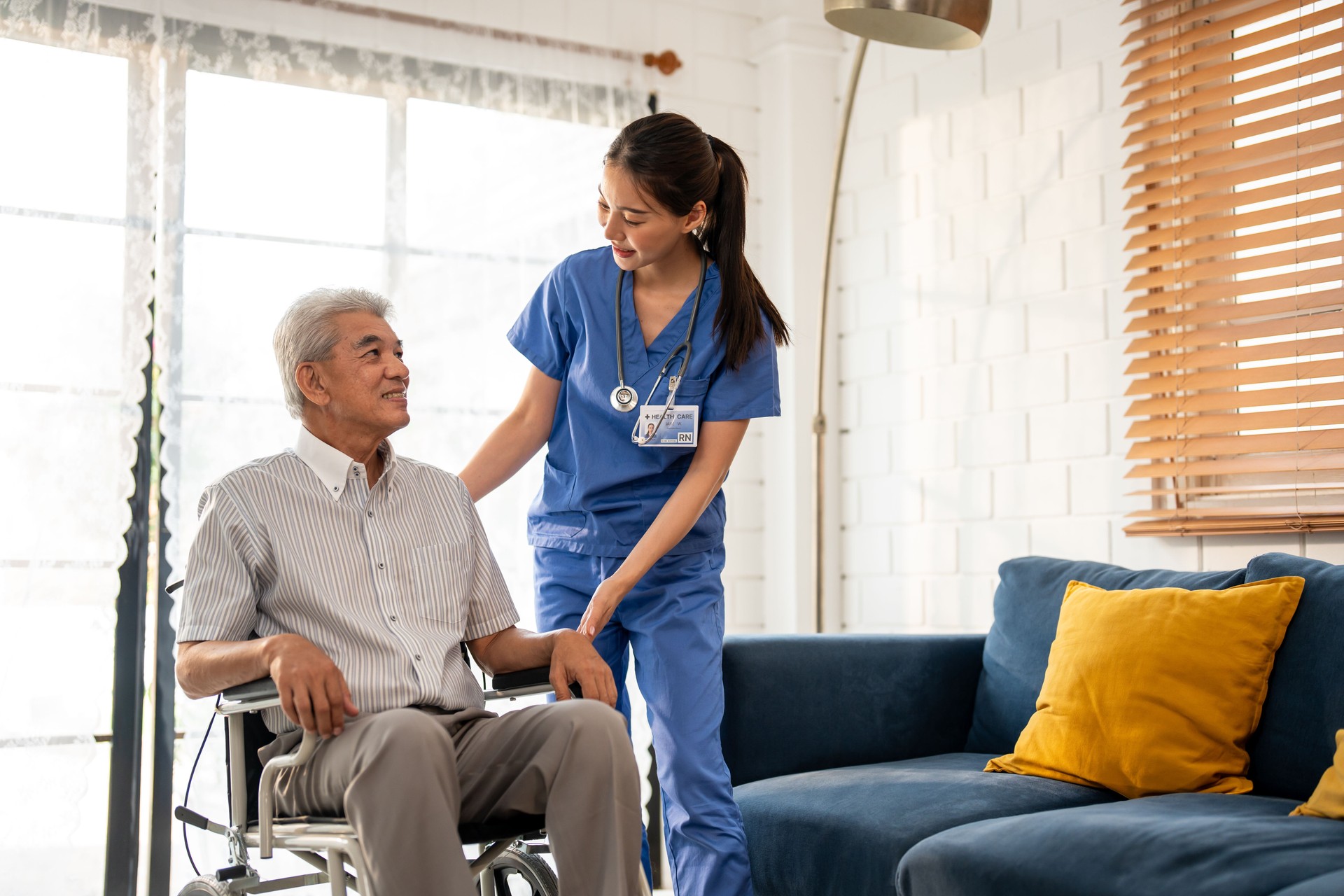Asian young caregiver take care senior man on wheelchair in house. Attractive nurse doctor support elderly older mature male patient sitting on sofa at nursing home care. Medical insurance concept