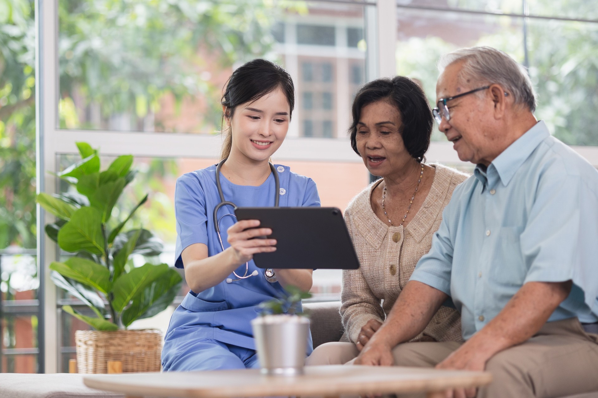 A smiling young nurse assists an elderly couple with technology, showing them information on a tablet in a bright, plant-filled room.